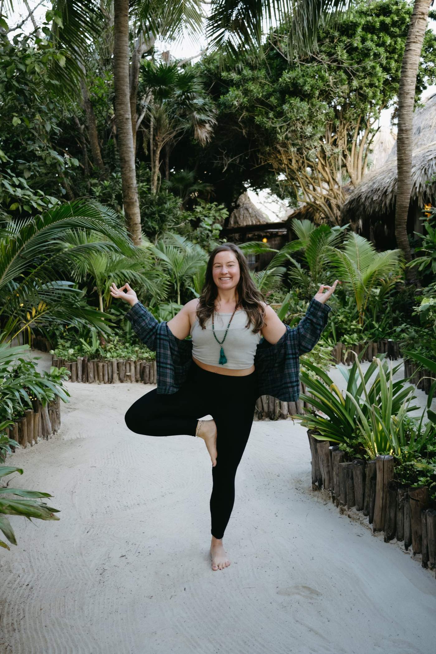 Megan smiling in tree pose on a sandy, palm-lined path